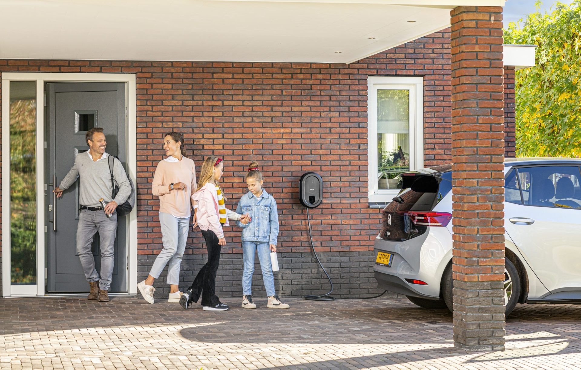 Gezin vertrekt vrolijk vanuit huis, met een elektrische auto aan de laadpaal onder de carport.