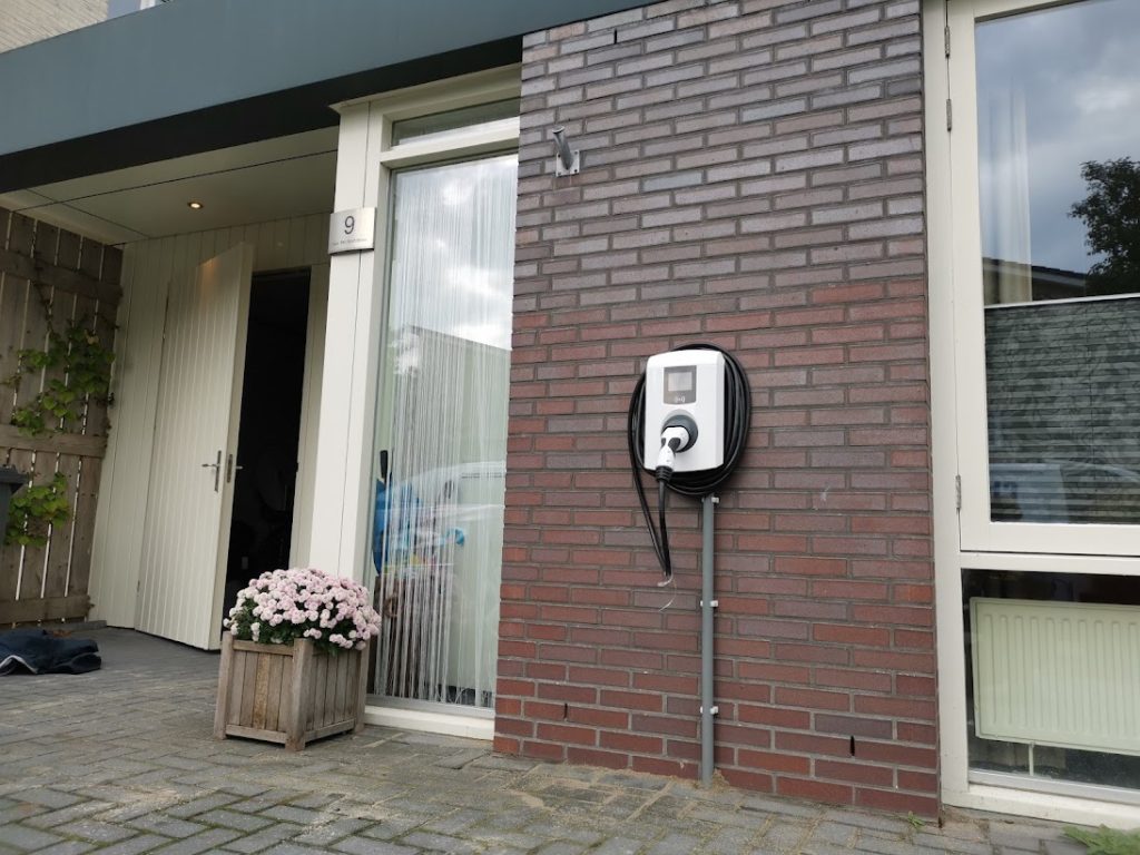 Wandgemonteerde laadpaal thuis met opgebonden kabel aan buitenmuur naast voordeur van woning (huisnummer 9), klaar voor elektrisch laden; professionele installatie door LaderThuis.nl.
