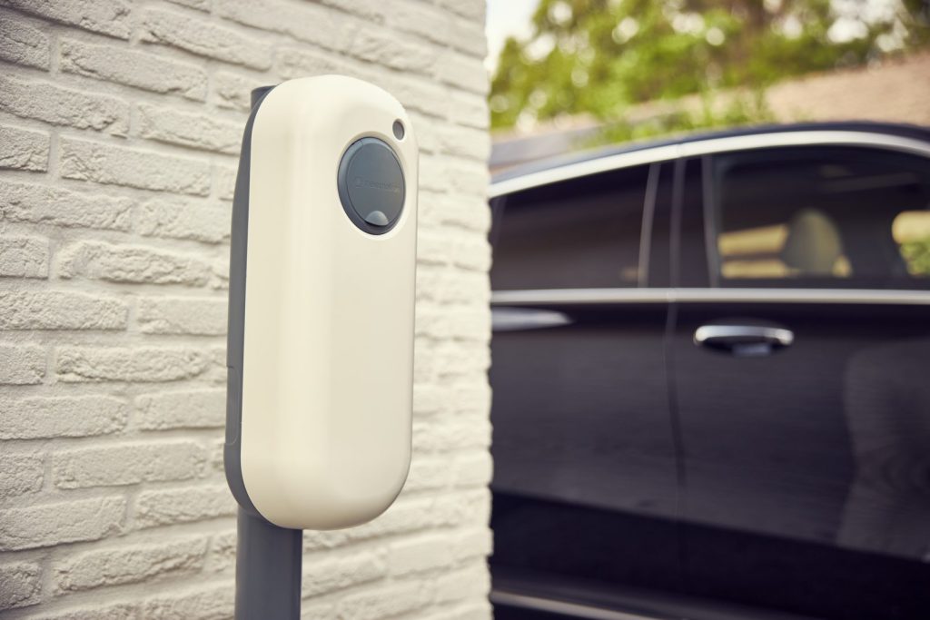 Witte NewMotion laadpaal op een paal voor een geparkeerde zwarte auto bij een wit gemetselde muur.