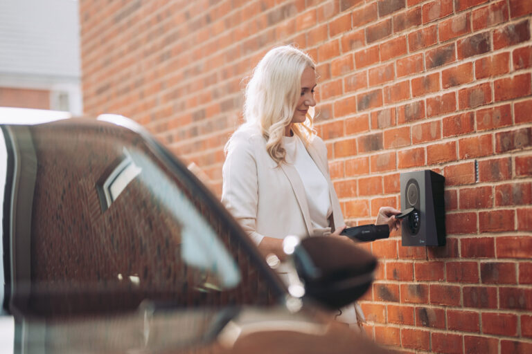 Vrouw sluit een elektrische auto aan op een zwarte Zaptec laadpaal aan een rode bakstenen muur.