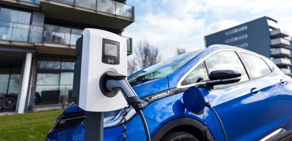 Blauwe elektrische auto laadt op bij een openbare laadpaal voor een modern appartementencomplex.