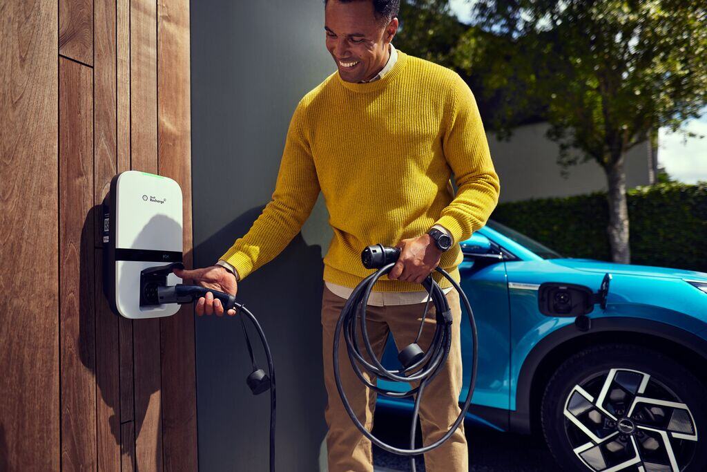 Man sluit laadsnoer aan op een laadstation aan een houten wand, met een elektrische auto op de achtergrond.