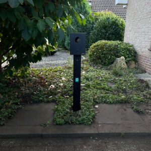 Zwarte vrijstaande laadpaal thuis op paal, geïnstalleerd naast tuinpad bij huis tussen groene struiken.