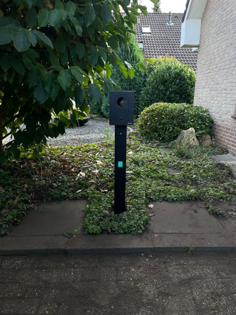 Zwarte vrijstaande laadpaal thuis op paal, geïnstalleerd naast tuinpad bij huis tussen groene struiken.