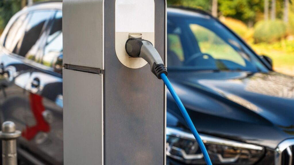 Elektrische laadpaal met aangesloten blauwe laadkabel laadt een zwarte auto op, geparkeerd op een buitenparkeerplaats met bomen en groen op de achtergrond.