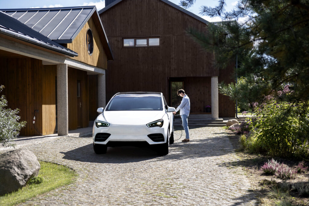 Elektrische auto wordt opgeladen terwijl een persoon een laadkabel aansluit aan de laadpaal thuis bij een houten woning met oprit.