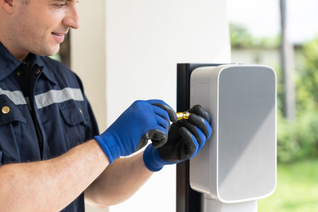 Wallbox aan buitenpaal, gecertificeerde monteur draait schroef vast tijdens laadpaal installatie thuis, blauwe werkhandschoenen zichtbaar, huis en tuin op de achtergrond.