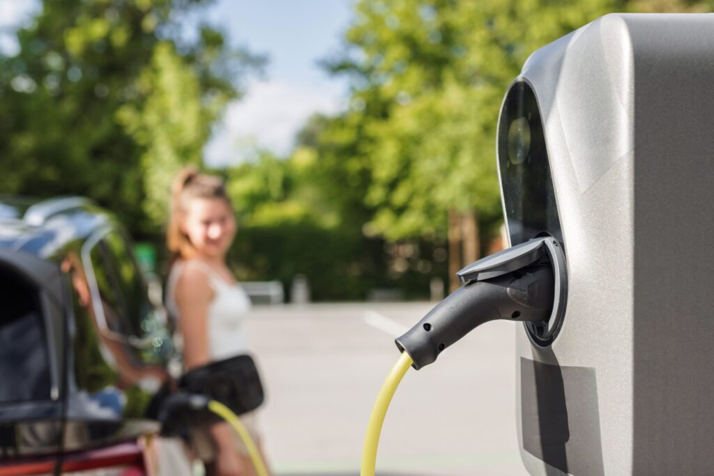 Laadkabel verbonden met laadpaal, laadt elektrische auto terwijl vrouw bij de geparkeerde auto staat op een zonnige parkeerplaats met groene bomen op de achtergrond.